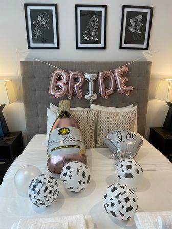 Bridal suite bedroom with tufted headboard and rose-gold "BRIDE" balloons above a bed, oversized champagne and "I do" balloons and black-and-white cow-print balloons scattered on white linens, flanked by bedside lamps and botanical wall art.