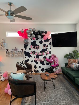 Home living room party setup with cow-print backdrop, “LET’S GO GIRLS” banner, pink, black and silver balloon garland, disco ball and pink cowboy boot balloon.