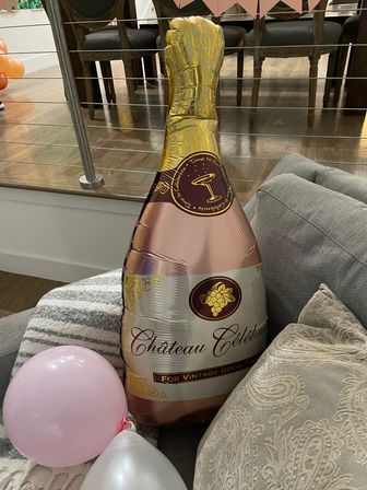 Rose-gold and gold foil champagne-bottle balloon on a sofa with pink and white balloons, indoor living-room party decor with wood floor and dining chairs in background.