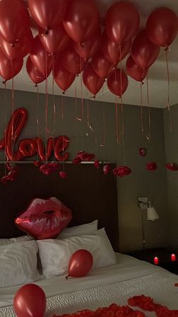 Romantic hotel room with red helium balloons, foil "love" and lips balloons, scattered rose petals and lit candles on the bed.