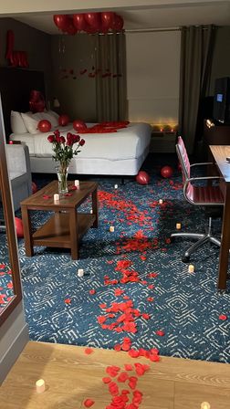 Romantic hotel room setup with red rose petals trailing across patterned carpet to a king bed, red balloons overhead, vase of red roses on a table and glowing LED candles.
