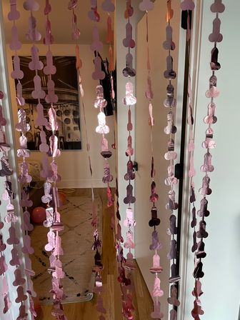 Playful pink metallic novelty garland of repeated phallic shapes draping a home doorway, framing a hallway with hardwood floors, a rug and scattered balloons.