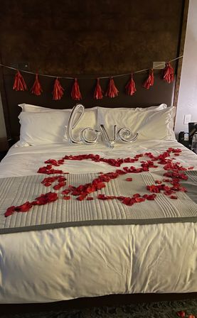 Romantic hotel bed with white linens, red tassel garland on the headboard, a silver 'love' balloon and red rose petals arranged in a heart on the comforter.