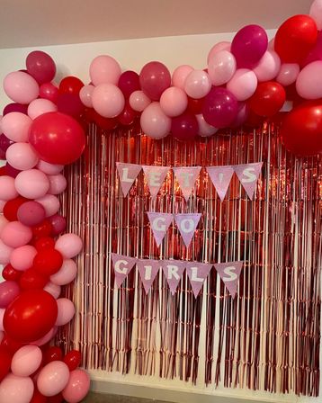 Festive party photo backdrop with pink and red balloon garland and rose-gold metallic fringe curtain, glitter triangular pennant banner reading 'LET'S GO GIRLS'.