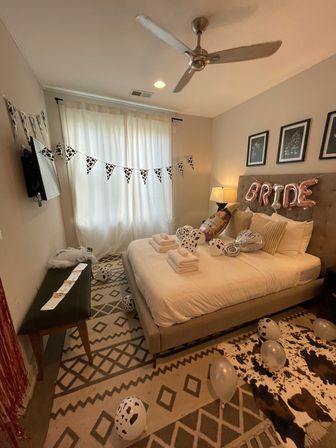 Cozy modern bedroom staged for a bridal celebration with a tufted bed topped by rose-gold "BRIDE" balloons, champagne and black-and-white spotted balloons, matching pennant bunting, neatly folded towels on the bed, neutral bedding, patterned rug and ceiling fan.
