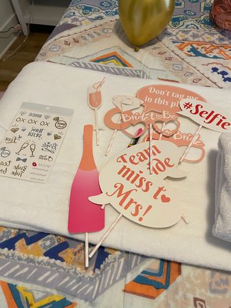 Bachelorette party photo booth props on a bed: pink champagne bottle and glass cutouts, speech bubbles reading “Team bride” and “miss to Mrs!”, #Selfie sign and a gold balloon on a colorful patterned bedspread.