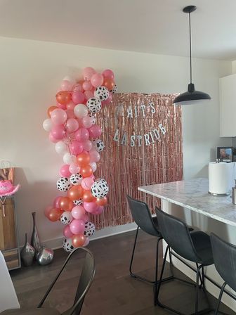 Home kitchen party setup with cascading pink, coral and black-spotted balloons framing a rose-gold fringe backdrop behind a marble island and black bar stools.