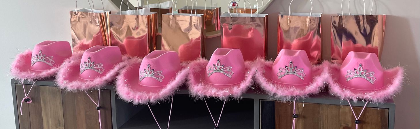 Row of pink feather-trimmed cowgirl hats with rhinestone tiaras lined up in front of rose-gold gift bags — playful party favors