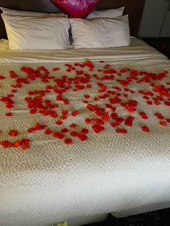 Romantic hotel bed with red rose petals scattered across a white quilt, two pillows and a pink heart balloon