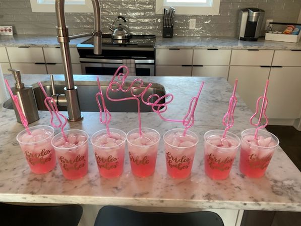 Eight pink iced cocktails in clear plastic cups with playful pink swirly straws arranged on a marble kitchen island in a modern white kitchen — bridal party drink setup.
