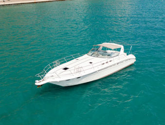 Sleek white motor yacht floating on bright turquoise water beside an urban waterfront promenade with trees and parked cars
