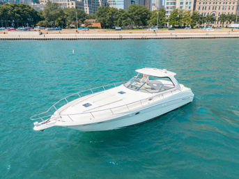 Aerial view of a white motor yacht cruising turquoise water near an urban tree-lined waterfront and city skyline on a sunny day.