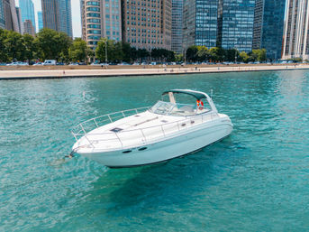White cabin motorboat on turquoise water near an urban waterfront with tree-lined promenade and high-rise buildings on a sunny day