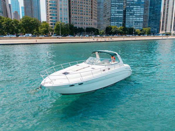 White cabin motorboat on turquoise water near an urban waterfront with tree-lined promenade and high-rise buildings on a sunny day