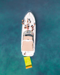 Aerial drone view of a white yacht with people sunbathing on the deck and a bright yellow-green inflatable mat trailing in clear turquoise water.