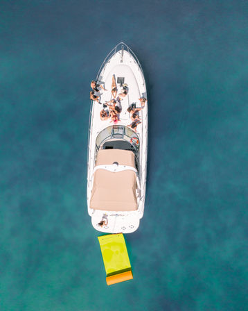 Aerial drone view of a white yacht with people sunbathing on the deck and a bright yellow-green inflatable mat trailing in clear turquoise water.