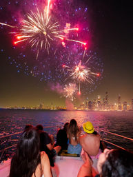 Friends lounging on a boat bow at night watching vibrant pink, red and blue fireworks burst over a glowing city skyline with shimmering reflections on the water, one person wearing a yellow hat.