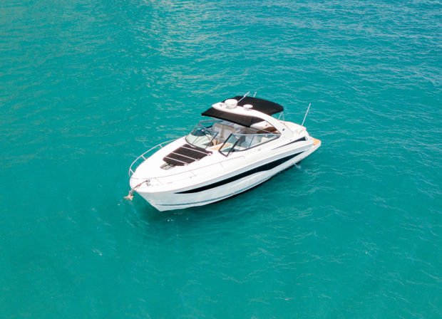 Sleek white motor yacht anchored in clear turquoise water near a sunlit coastal promenade with parked cars and pedestrians