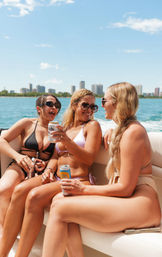 Three friends in bikinis laughing and holding canned drinks while lounging on a boat with blue water and an urban skyline under a sunny summer sky.