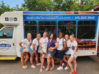 Seven women in casual summer outfits and matching shirts pose and laugh in front of a Nashville tour shuttle decorated with a city skyline graphic, parked by trees.