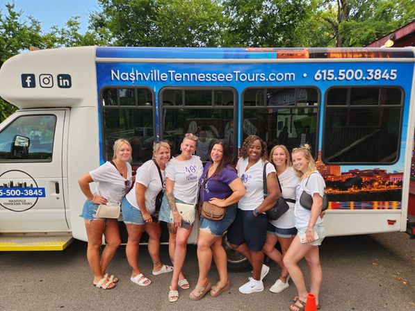 Seven women in casual summer outfits and matching shirts pose and laugh in front of a Nashville tour shuttle decorated with a city skyline graphic, parked by trees.