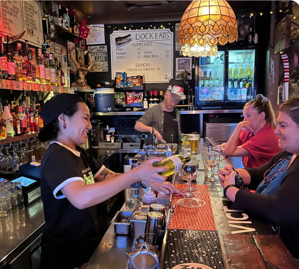 Friendly bartender pouring white wine for smiling patrons at a cozy waterfront bar counter, warm pendant lamp, stocked liquor shelves, menu board and beer taps in the background.