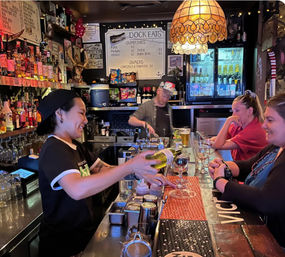 Friendly bartender pouring white wine for smiling patrons at a cozy waterfront bar counter, warm pendant lamp, stocked liquor shelves, menu board and beer taps in the background.