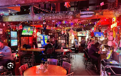 Neon-lit cozy dive bar interior with wooden tables and high-top stools, colorful string lights and holiday decorations, slot machines and TV screens, patrons seated at the bar and gaming machines in a lively neighborhood pub atmosphere.