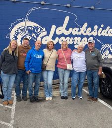 Seven people smiling arm‑in‑arm in a parking lot, posing in front of a bright blue wall mural with a white stylized fish and large script lettering; casual jeans and jackets in an outdoor daytime group photo.