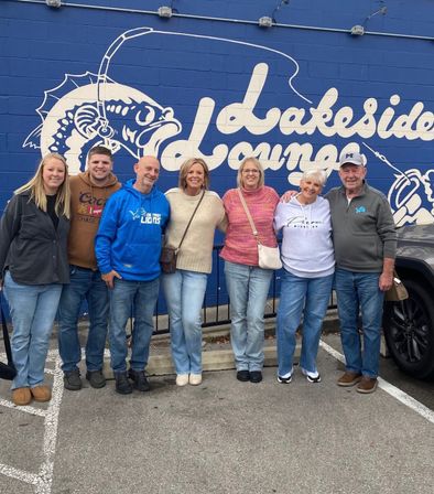 Seven people smiling arm‑in‑arm in a parking lot, posing in front of a bright blue wall mural with a white stylized fish and large script lettering; casual jeans and jackets in an outdoor daytime group photo.