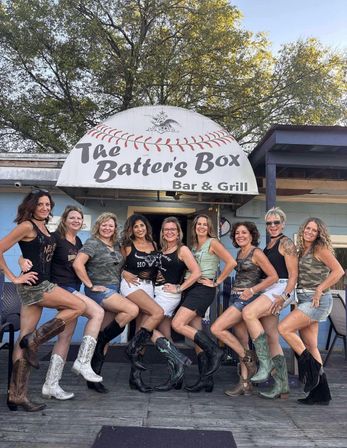 Ten women smiling and kicking heels together on a wooden deck outside a bar and grill, wearing denim shorts, camo and tank tops and colorful cowboy boots beneath leafy trees on a sunny afternoon.