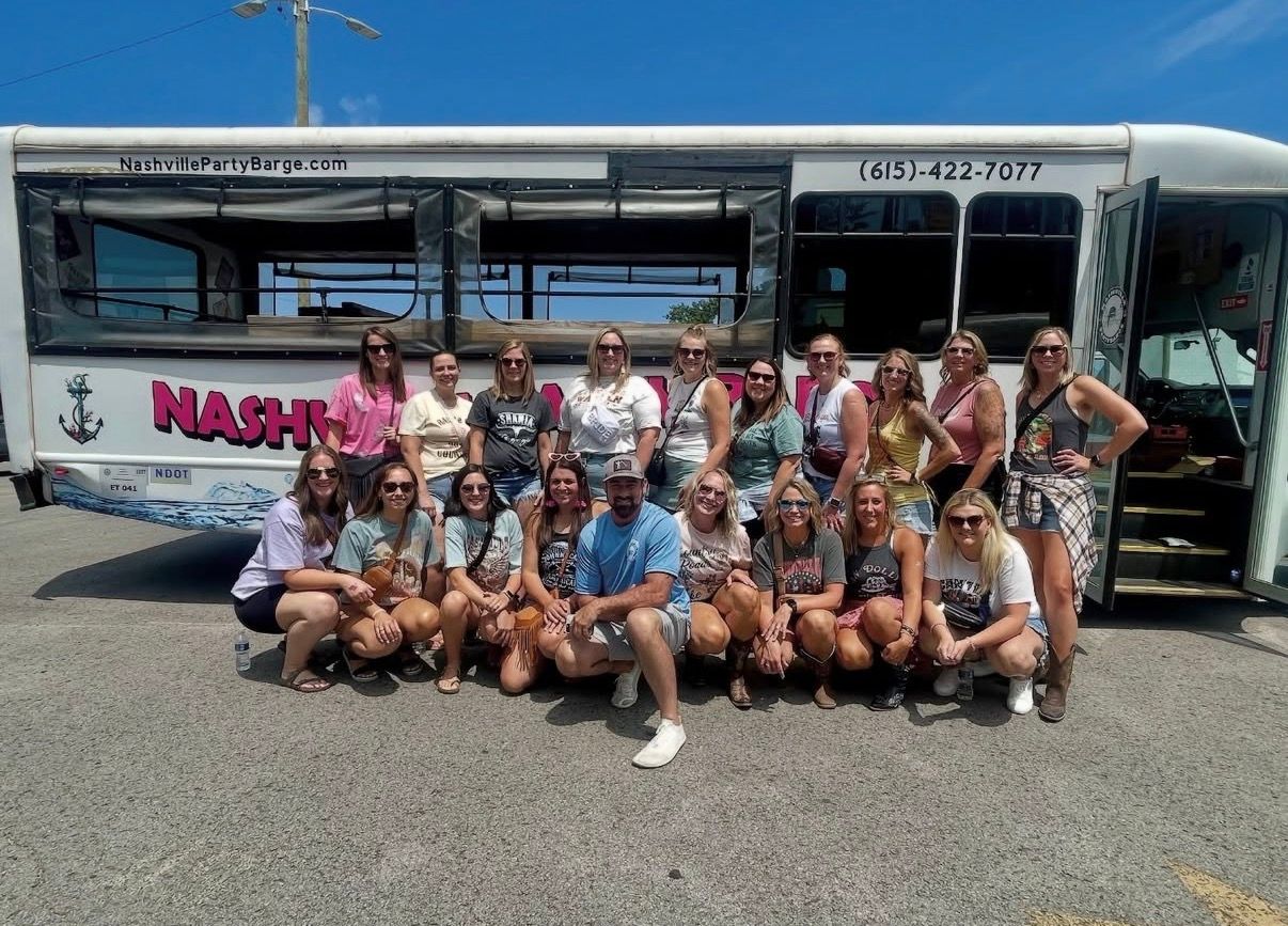 Group of about 20 adults smiling and posing in front of a white party bus with pink lettering in Nashville on a sunny day.