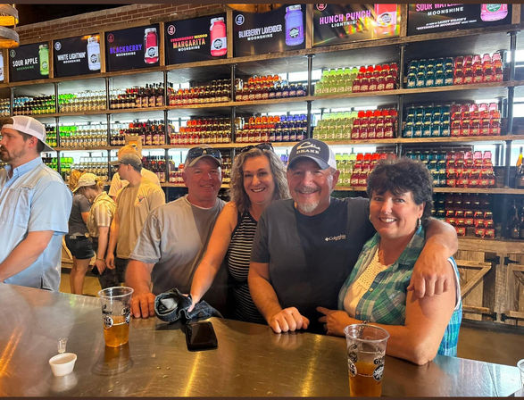 Four smiling adults leaning on a metal bar in an indoor distillery tasting room, colorful rows of bottled moonshine and flavored jars on wooden shelves behind them, with plastic cups of beer on the counter.
