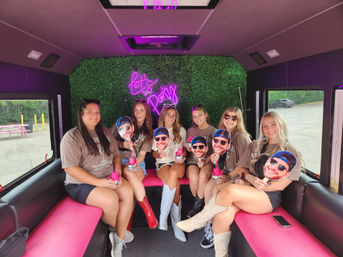 Seven friends smiling inside a pink-and-black party bus with a neon "Let's Party" sign and faux-boxwood wall, holding novelty face cutouts and pink drink cups while seated on bright bench seats.