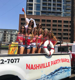 Bachelorette group of women in matching red tanks and sunglasses partying on a daytime Nashville river barge, bride in a veil holding up a drink with city high-rises in the background.