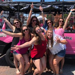 Seven friends in summer outfits laughing and cheering in front of a pink party vehicle on a sunny downtown street — playful bachelorette-style celebration.