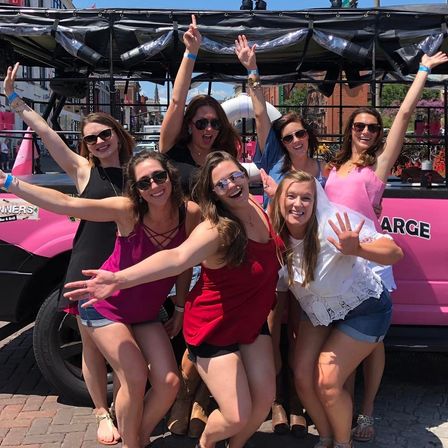 Seven friends in summer outfits laughing and cheering in front of a pink party vehicle on a sunny downtown street — playful bachelorette-style celebration.