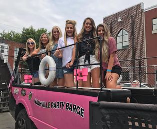 Six smiling women on a pink open-top party barge vehicle parked on a downtown Nashville street, ready to celebrate