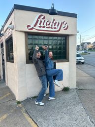 Two friends in denim and sneakers striking a playful pose outside a corner neighborhood bar with a vintage painted sign and green glass-block window on an urban street.
