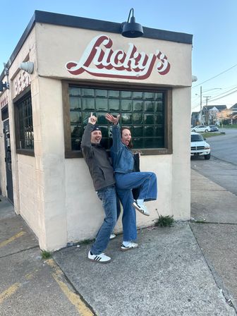 Two friends in denim and sneakers striking a playful pose outside a corner neighborhood bar with a vintage painted sign and green glass-block window on an urban street.