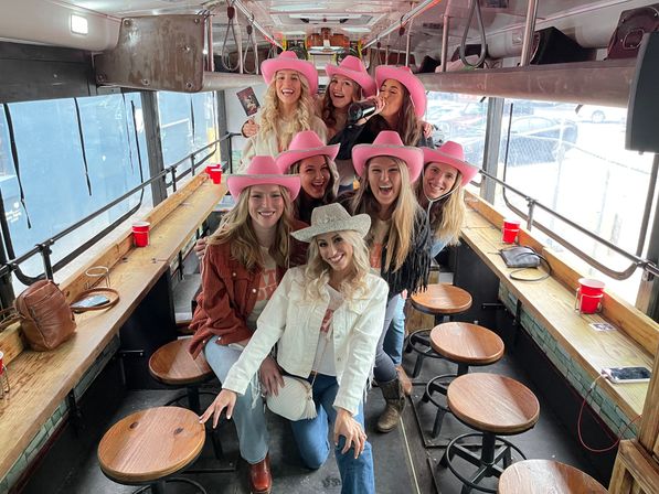 Group of women in pink cowboy hats laughing and posing on a party bus with wooden bar seating, round stools, red solo cups and a daytime parking lot visible through the windows — lively celebration vibe.