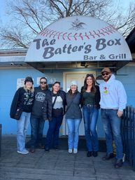 Six people posing in a group photo on a wooden deck outside a blue bar-and-grill entrance with a large baseball-themed awning, wearing winter coats, hats and jeans beneath bare winter trees.
