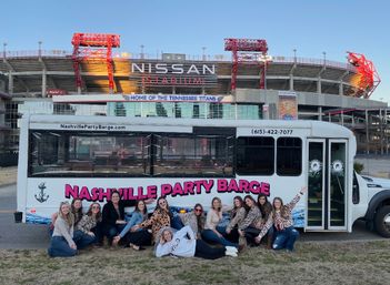 Group of friends posing in front of a white party-barge bus with pink lettering, parked outside a large football stadium in Nashville, Tennessee at dusk