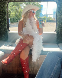 Bride-to-be in a white cowboy hat and feather boa, heart-shaped sunglasses and red star red cowboy boots, holding a canned drink while seated in a sunlit shuttle with greenery-lined interior and city skyline visible through the window.