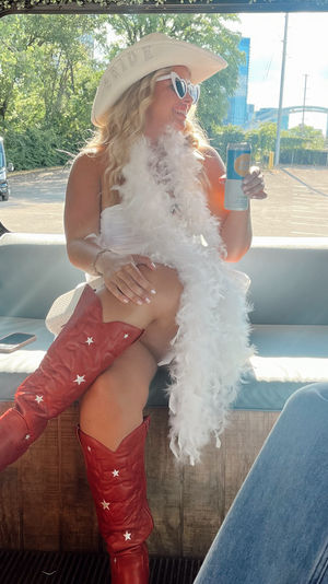 Bachelorette in white dress and feather boa wearing a 'BRIDE' cowboy hat and heart sunglasses, sipping a drink aboard a party bus with red star cowboy boots and city skyline outside