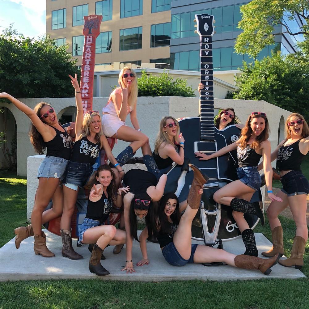 Bachelorette crew in denim shorts, cowboy boots and sunglasses laughing and striking playful poses on oversized decorative guitar sculptures in a sunny outdoor plaza