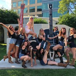 Bachelorette crew in denim shorts, cowboy boots and sunglasses laughing and striking playful poses on oversized decorative guitar sculptures in a sunny outdoor plaza