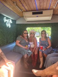 Three friends in sunglasses and cowboy boots sitting inside a decorated party bus with faux greenery walls, a neon "Good Vibes Only" sign, wooden ceiling with blue LED lights and an AC unit, holding drinks with a sunny green view through the window.