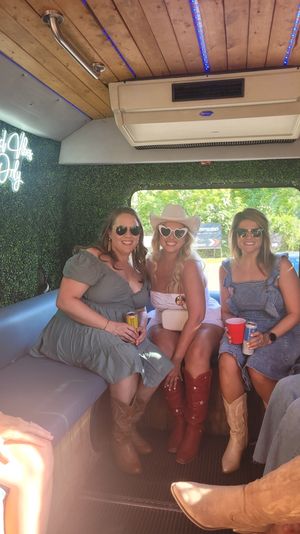 Three friends in sunglasses and cowboy boots sitting inside a decorated party bus with faux greenery walls, a neon "Good Vibes Only" sign, wooden ceiling with blue LED lights and an AC unit, holding drinks with a sunny green view through the window.