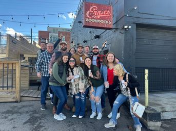 Group of friends posing outside an urban bar with string lights and a wooden patio, smiling and holding drinks on a sunny day
